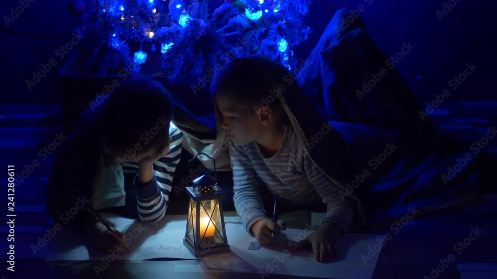 Funny children write letter with pictures of Santa Claus under fir, covered with blanket and flashlight. Children are happy to spend winter holidays together. Concept of traditional family holidays. Funny children write letter with pictures of Santa Claus under fir, covered with blanket and flashlight. Children are happy to spend winter holidays together. Concept of traditional family holidays.