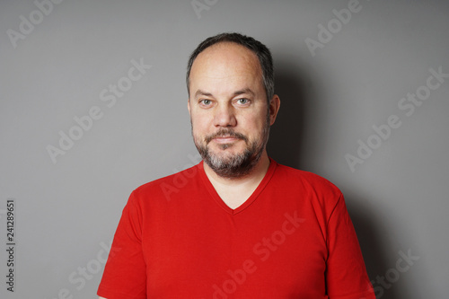 middle aged man in his 40s wearing red t-shirt with short dark hair and graying beard smirking - gray wall background with copy space photo