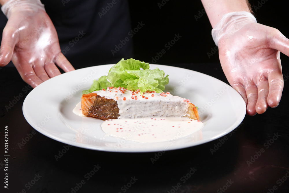 The hands of the chef serving dishes from a piece of salmon in a creamy caviar sauce on a white plate with salad leaves. Macro photo.