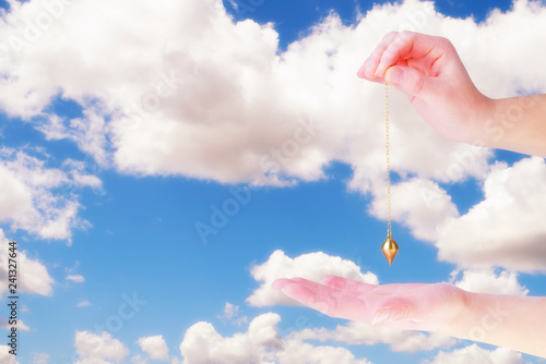 Close up of woman hand holding a pendulum over her palm. Blue sky and white clouds in background.