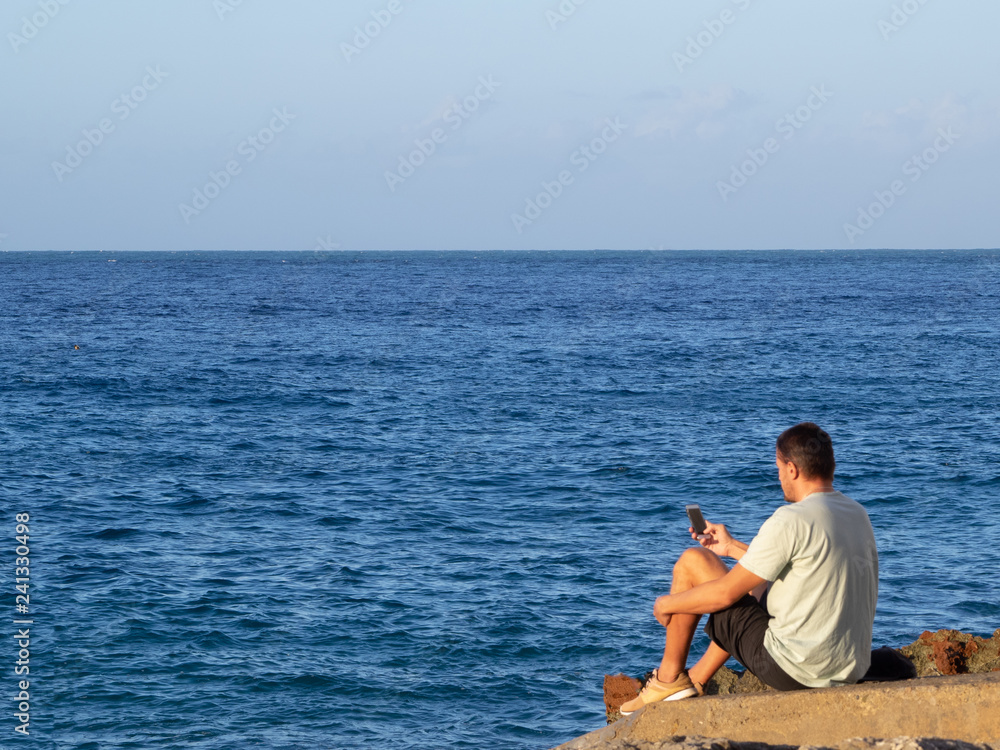 adult man on vacation on the coast of the blue sea in summer clothes with a smartphone in his hand.