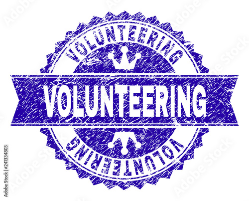 VOLUNTEERING rosette seal watermark with grunge style. Designed with round rosette, ribbon and small crowns. Blue vector rubber watermark of VOLUNTEERING label with retro style.
