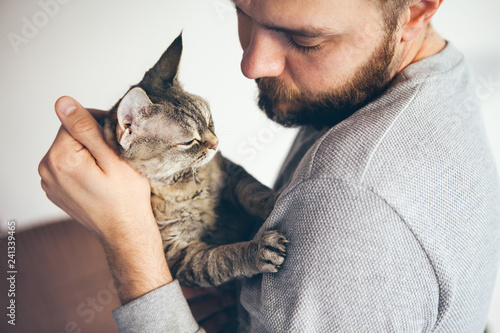 Close-up of beard man hugging cuddly cat, lifestyle portrait. Caring man is holding a Devon Rex -tabby kitty, both looking at each other with affection. Lifestyle, home interior photo