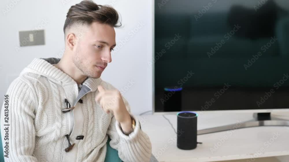 Young handsome man speaking to smart electronic speaker home assistant, sitting on couch in living-room Young handsome man speaking to smart electronic speaker home assistant, sitting on couch in living-room