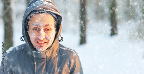 Frozen young man in a jacket with a hood covered with snow in winter forest. Snowflakes lie on the eyelashes, eyebrows, cheeks. photo