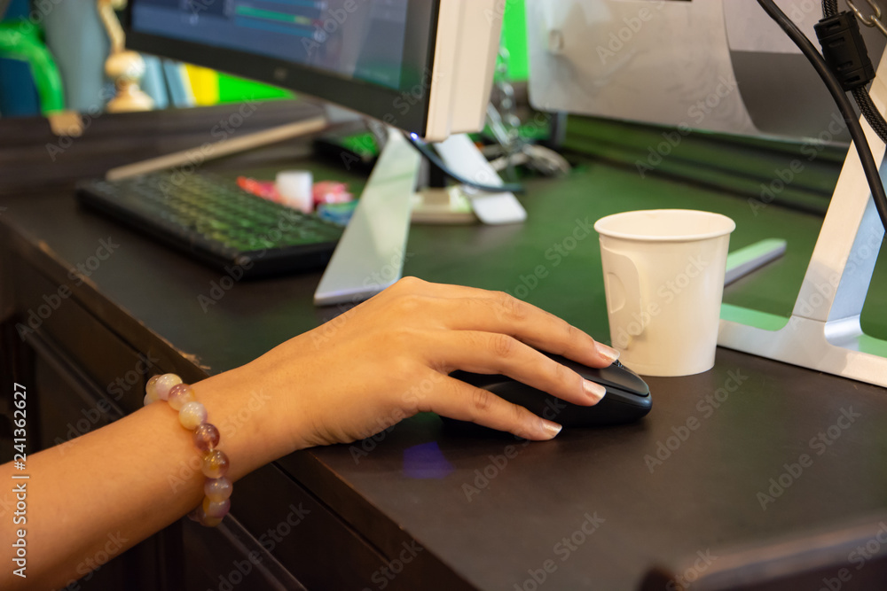 The young woman's hand is working, holding the black mouse on the desk in front of the computer. The young woman's hand is working, holding the black mouse on the desk in front of the computer.