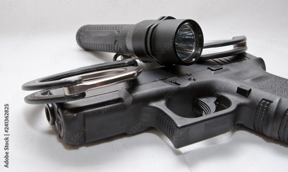A pair of black and silver handcuffs with a black flashlight laying on top of a black semi automatic pistol on a white background A pair of black and silver handcuffs with a black flashlight laying on top of a black semi automatic pistol on a white background