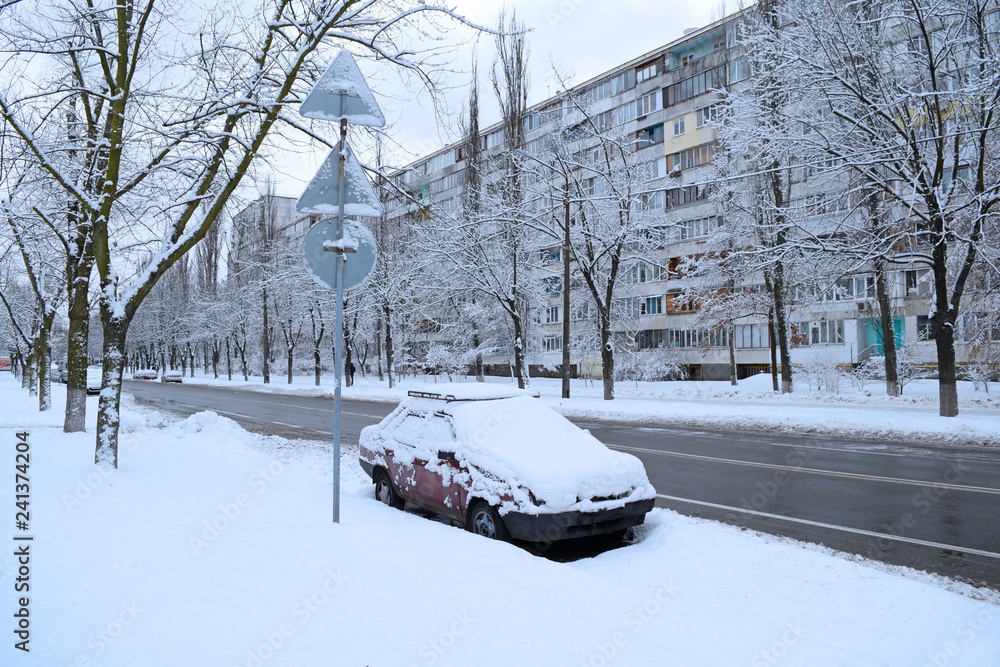 A small car in the snow stands near the asphalt road in the city in winter. A small car in the snow stands near the asphalt road in the city in winter.