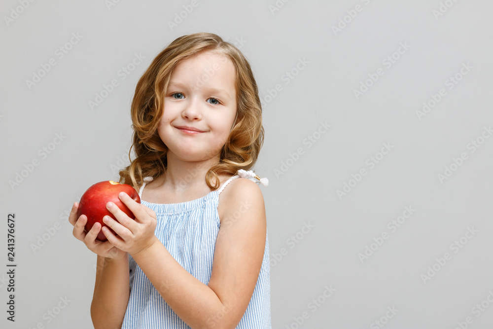 Portrait of a happy smiling little blonde girl on a gray background. Baby eating red apple Portrait of a happy smiling little blonde girl on a gray background. Baby eating red apple
