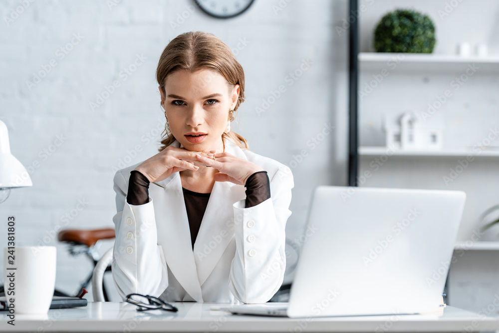 businesswoman in white formal wear with folded hands sitting at computer desk while looking at camera businesswoman in white formal wear with folded hands sitting at computer desk while looking at camera