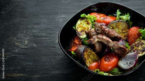 Baked beef and grilled vegetables in a black plate. On a wooden background. Top view. Free space for your text.