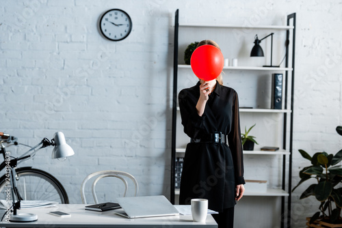 businesswoman in black clothes standing near table and hiding face behind red balloon