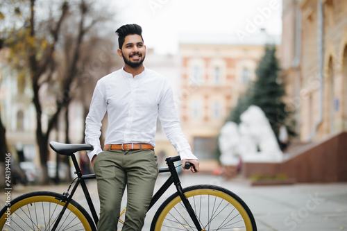 Fototapeta Naklejka Na Ścianę i Meble -  Happy young stylish indian man dressed in shirt standing with a fixed gear bicycle