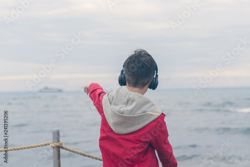 A boy in a red jacket and headphones looks at the sea waves in stormy rainy weather