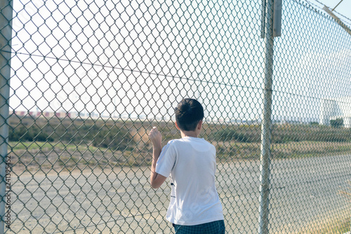 A barefoot boy stands on the beach near the iron net and looks at the airport.