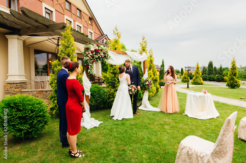 newlyweds standing face to face near decorated arch and witnesses newlyweds stand behind them. The master of ceremonies speaks to the microphone in the yard of the restaurant