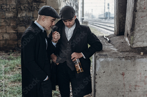 stylish gangsters men, smoking. posing on background of railway with bottle of alcohol. england in 1920s theme. fashionable brutal confident group. atmospheric  moments