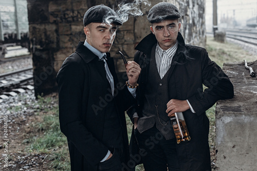 stylish gangsters men, smoking. posing on background of railway with bottle of alcohol.sherlock holmes and doctor watson. england in 1920s theme.