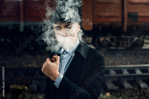 stylish gangster smoking in tweed look, posing on background of railway. england in 1920s theme. fashionable brutal confident man. atmospheric smoky moments. space for text