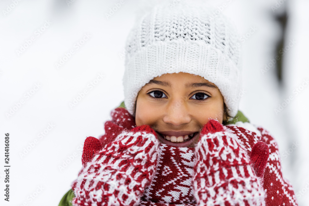 close up view of african american child in knitted hat, mittens and scarf smiling and looking at camera in winter close up view of african american child in knitted hat, mittens and scarf smiling and looking at camera in winter