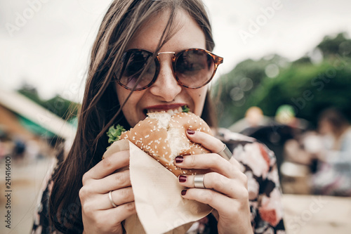 stylish hipster woman eating juicy burger. boho girl biting yummy cheeseburger, smiling at street food festival. summertime. summer vacation travel picnic. space for text photo