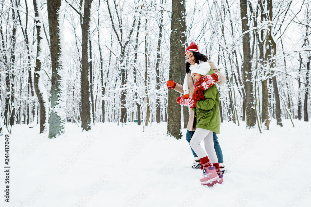 attractive african american mother and cute daughter walking and holding cups in snowy forest