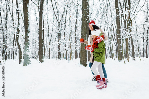 attractive african american mother and cute daughter walking and holding cups in snowy forest