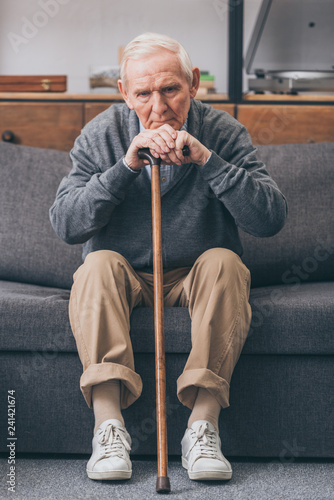 upset retired man with grey hair holding walking cane in living room