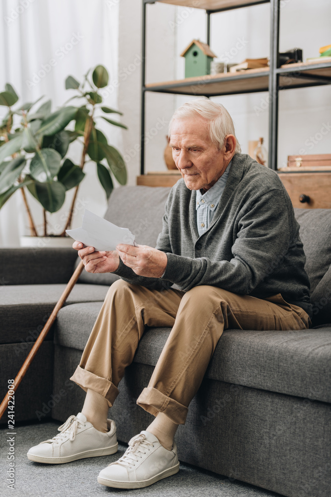 sad pensioner with grey hair looking at photos while sitting on sofa