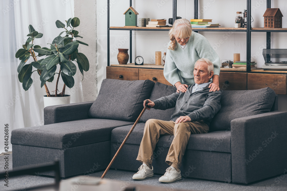 Obraz premium retired wife embrace sad senior husband sitting with walking cane in living room