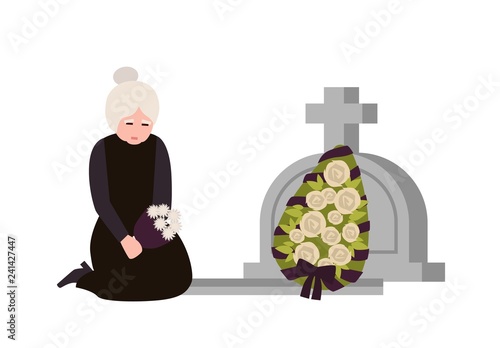 Sorrowful elderly woman dressed in mourning clothes crying near grave with headstone and wreath