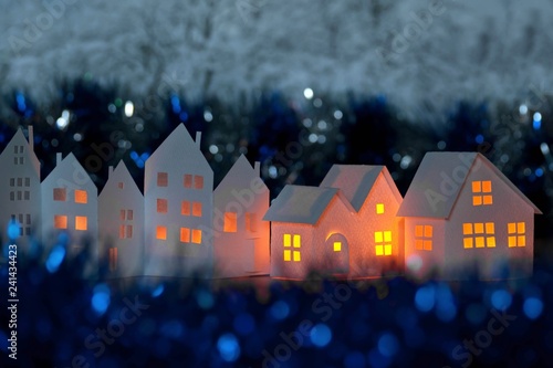 Handmade small white folded cardboard houses with illuminated windows on dark blue bokeh background. Winter decoration, evening dusk. Architecture business real estates concept. Shallow depth of focus