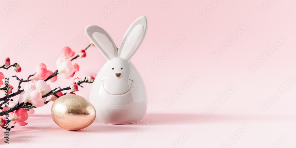 Obraz premium Easter golden eggs, pink flowers and porcelain rabbit on pastel pink background. Easter, spring concept. Copy space, banner