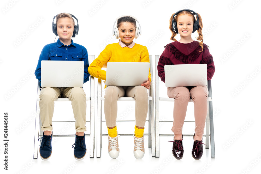 Obraz premium smiling children with headphones sitting on chairs, using laptops and looking at camera isolated on white