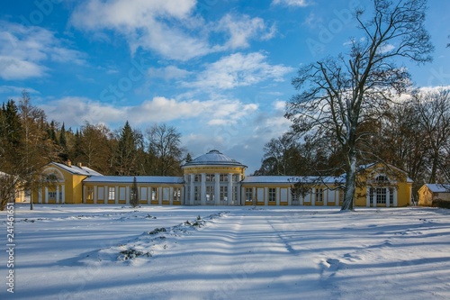 Winter scene of yellow building of Ferdinand colonnade with mineral water at spa town Marienbad, Czech Republic, park with trees, ground covered with snow, sunny day, blue sky, white clouds © Lioneska