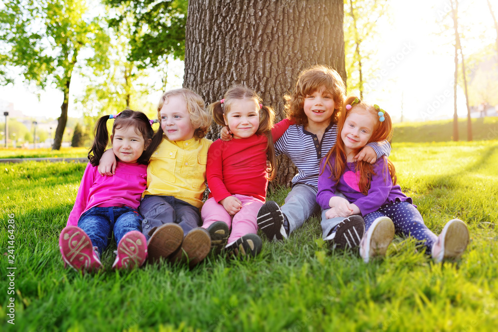 group of happy little children smiling sitting in park on grass under a tree. June 1, Children's Day, friendship, childhood, vacation. group of happy little children smiling sitting in park on grass under a tree. June 1, Children's Day, friendship, childhood, vacation.
