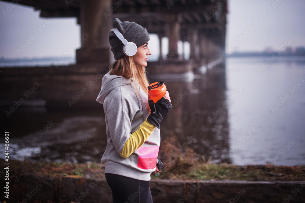 Woman drinking a protein shake in headphones. Young woman resting after running in winter