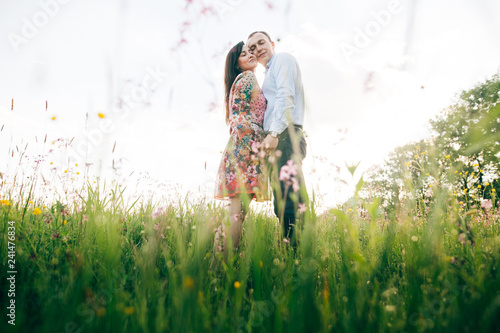 Beautiful young couple gently hugging  in sunshine in fresh spring meadow with pink flowers. Happy stylish family embracing in green field in sunlight. Romantic moments. Hello Spring