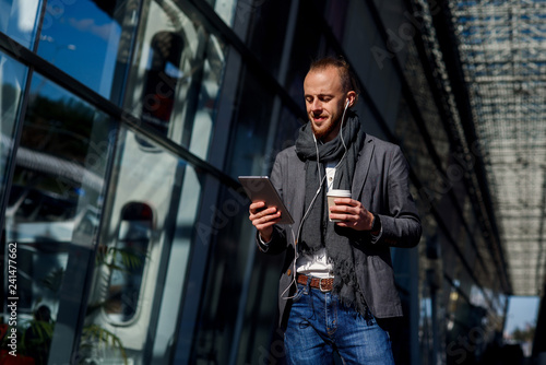 Attractive caucasian businessman wearing casual clothes walking outside, holds tablet computer and drinks coffee near modern office centre. Business concept.
