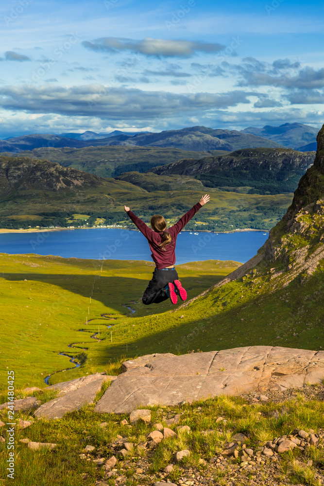 Young Pretty Girl Jumps Happily From Rock In Front Of Spectacular Landscape Of Applecross Pass In Scotland