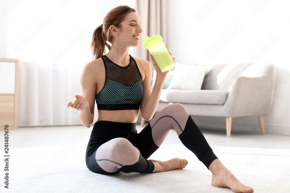 Young sporty woman with bottle of protein shake sitting on floor at home
