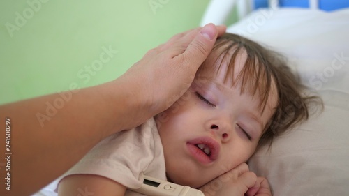mom measures temperature with thermometer to baby and caresses child with hand. close-up. baby sleeps in hospital ward on white bed.