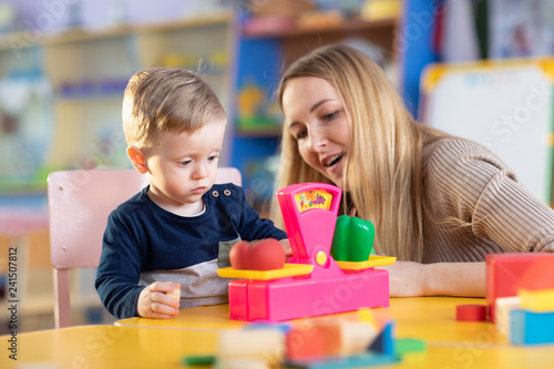 Little child boy and nursery teacher sit at table and play with toy scales in playroom