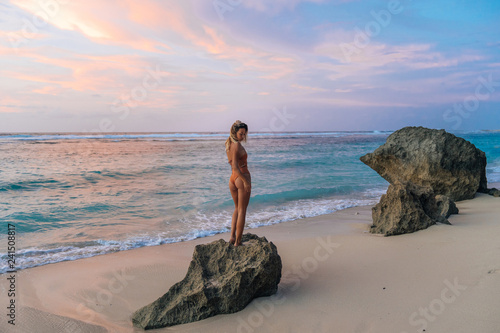 Woman with perfect tanned body is standing on big stone on beach during beautiful sunset