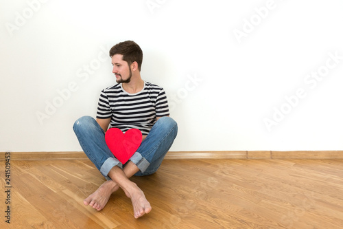 Happy valentines day! Young man is sitting on the floor with red heart and smiling. Copy space.