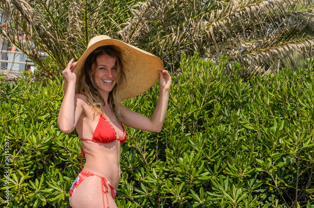 Young charming girl in a big straw hat stands among tropical palm trees in a swimsuit on a sunny summer day