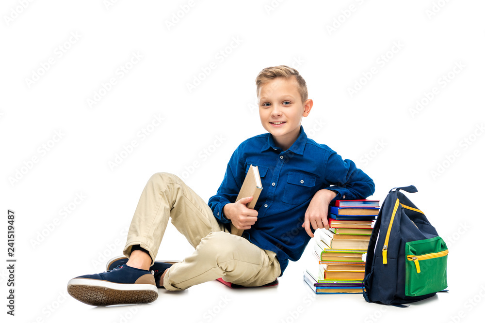 smiling schoolboy holding book, looking at camera and sitting near backpack and stack of books isolated on white smiling schoolboy holding book, looking at camera and sitting near backpack and stack of books isolated on white