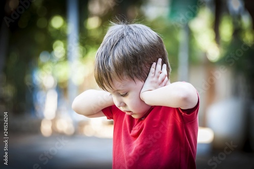 A 4 year old autistic child in a red shirt closing his ears with hands as if protecting from noise. Autism concept, asperger syndrome, loud noise, scared little kid, parents divorce trauma photo
