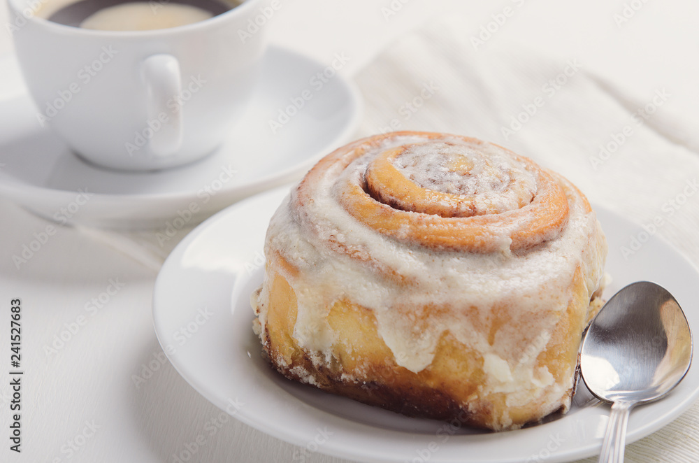 Hot cinnamon bun with sugar creamy icing on white plate. Sweet breakfast or snack with a cup of coffee.