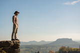 Young bearded male traveler in hat stand on the cliff in sunny day 